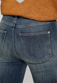 Jeans de mezclilla azul oscuro con un corte ajustado, diseño de cinco bolsillos y sutiles detalles de desgaste en la tela con costuras visibles.