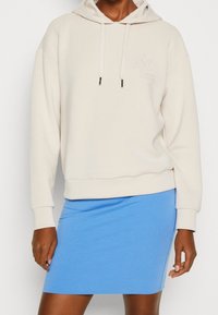 Sudadera beige con capucha, cordones y logotipo, combinada con una falda ajustada de color azul claro. Tela suave, diseño casual y estilo sencillo.