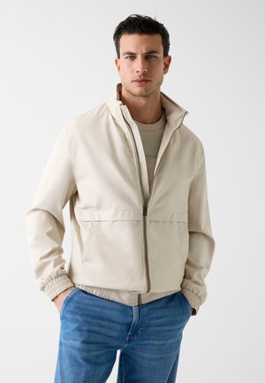 Junger Mann mit dunklen Haaren, der eine beige Reißverschlussjacke über einem hellen Pullover trägt und die Hände in den Taschen einer blauen Jeans hat.