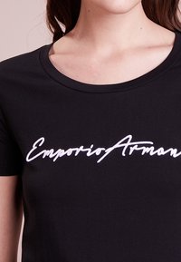 Camiseta de algodón negra con escote redondeado, que presenta un logo bordado en blanco "Emporio Armani" en el pecho. Mangas cortas.