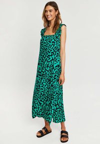 Grøn leopardprint maxi-kjole med flæse-stropper, firkantet udskæring og en bølgende silhuet. Kombineret med sorte slidesandaler.