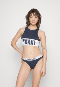 Marinblå bikini-set bestående av en höghalsskjorta med en vit rand som visar texten "TOMMY" och matchande högmidjade underdelar med logotryck på midjan.