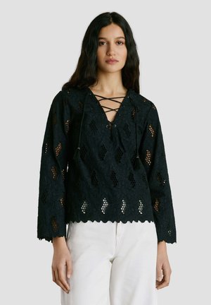 Femme aux longs cheveux bruns portant un haut en dentelle noire à manches longues avec un décolleté croisé et un pantalon blanc, sur un fond gris clair.