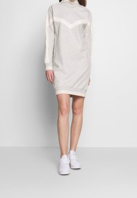 Graues Sweatshirtkleid mit hohem Kragen, kontrastierendem cremefarbenem Zickzackmuster, gerippten Bündchen und Saum. Getragen mit weißen Sneakern.