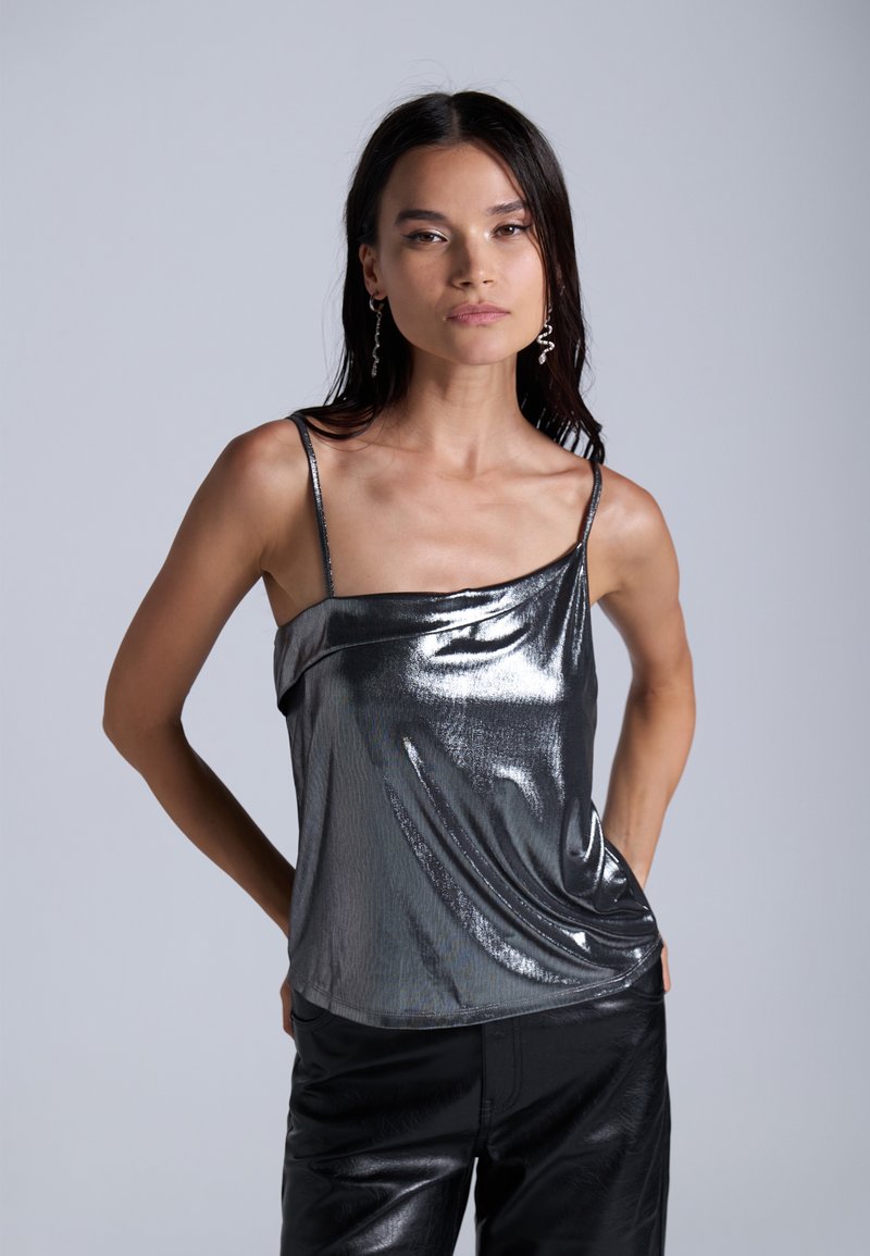 Metallic harmaa toppi, jossa on roikkuva muotoilu ja ohuet olkaimet, yhdistettynä korkeavyötäröisiin mustiin housuihin, joissa on sileä pinta ja kiilto.