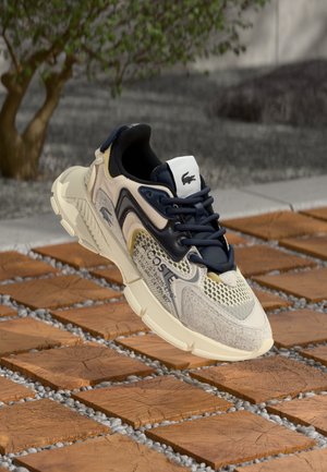 Lacoste Sneakers mit einem leichten Design in Creme und Marineblau, ausgestattet mit Mesh-Details, strukturierten Paneelen und einer Gummisohle, die über Holzfliesen schwebt.