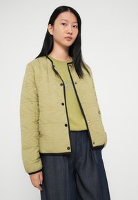 Quiltet olivengrøn jakke med sort kant, trykknaplukninger og en afslappet pasform, båret over en matchende grøn sweatshirt og brede mørke denimbukser.