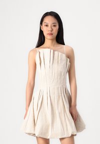 Une robe plissée beige clair avec des fines bretelles, dotée d'un tissu texturé et d'un corsage ajusté qui s'élargit en une jupe plus ample.