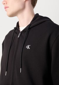 Sudadera con cremallera negra hecha de tejido suave, con un bolsillo canguro, cordones ajustables y un logo bordado en blanco en el pecho.