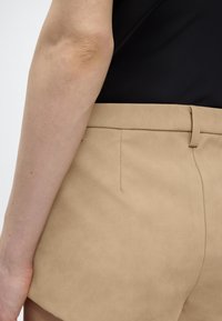 Κοντινό πλάνο του χεριού ενός ατόμου δίπλα σε μπεζ σορτς με ψηλή μέση, θηλιές για ζώνη και μαύρο τοπ που έχει μπει μέσα.