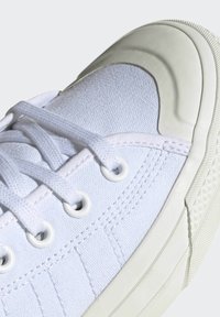 Zapatillas de lona blancas con puntera de goma, ojales y cordones blancos, suela texturizada y detalles cosidos en los laterales.