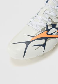 Botas de fútbol blancas con material sintético texturizado, con acentos de diseño en naranja y marino, cordones y una suela transparente con tacos.