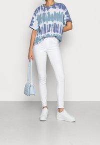 Person som bär en blå och vit tie-dye skjorta, vita skinny jeans, vita sneakers och håller i en ljusblå kviltad handväska.