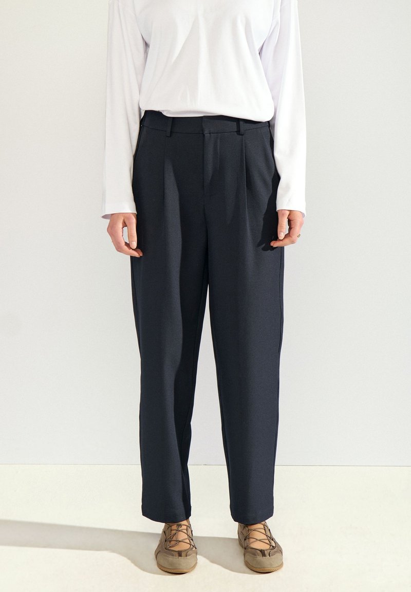 Personne portant un pantalon noir droit, un haut blanc à manches longues et des chaussures beiges à lacets, debout devant un fond uni clair.