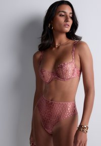 Ensemble de lingerie en dentelle rose comprenant un soutien-gorge balconnet avec un motif overlay et un shorty taille haute. Texture douce avec des accents métalliques.