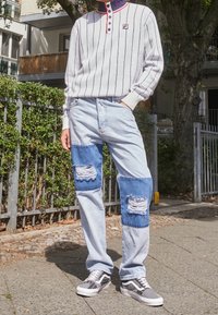 Camisa de mangas largas a rayas grises y blancas con acentos rojos, combinada con jeans de color azul claro que tienen parches de denim azul y bordes deshilachados. Zapatillas negras y blancas.