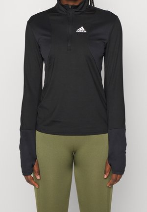Czarna koszulka Adidas z długim rękawem i ćwiartkowym zamkiem błyskawicznym z otworami na kciuki, noszona z oliwkowozielonymi legginsami na stojącej osobie.