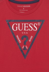 Rotes Baumwoll-T-Shirt mit einem graphischen navy-blauen Dreieck, auf dem der Text "GUESS", ein Fragezeichen und Produktdetails in Weiß und Navy gedruckt sind.