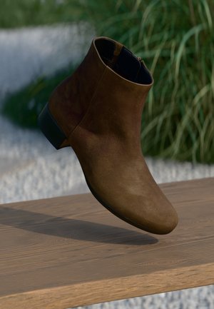 Stivale alla caviglia in suede marrone con punta arrotondata, cerniera laterale e tacco corto a blocco, posizionato su uno sfondo naturale.