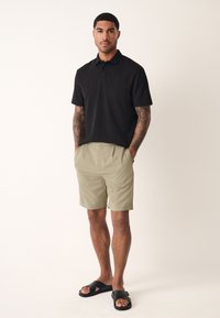 Polo noir à manches courtes texturé, short beige et sandales noires, coupe décontractée avec des détails subtils.