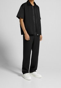 Czarna kurtka z krótkim rękawem na zamek, o swobodnym kroju, zestawiona z dopasowanymi czarnymi spodniami. Outfit dopełniają białe trampki ze złotymi akcentami.