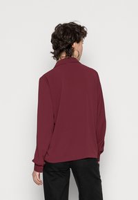 Langarmige bordeauxblouse met een kraag, vloeiende stof, knoopmanchetten en een ontspannen pasvorm, gecombineerd met zwarte broek.