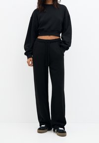 Schwarzer, verkürzter Sweatshirt mit überschnittenen Schultern, kombiniert mit schwarzen Jogginghosen mit elastischem Bund und Seitentaschen, getragen mit schwarzen Sneakern.