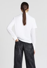Camicia bianca a maniche lunghe con maniche corte arrotolate, abbinata a jeans a gamba larga grigio scuro con tasche posteriori e una finitura testurizzata.
