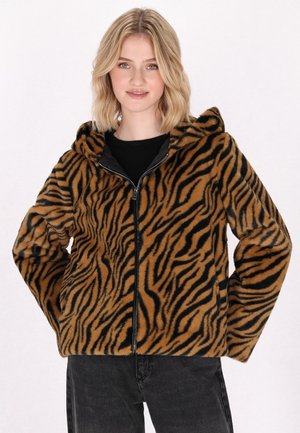 Junge Frau mit kurzen blonden Haaren trägt eine braun-schwarze tigergetigerte Reißverschlussjacke mit Kapuze und schwarze Hose.