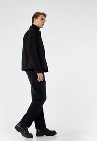 Chaqueta negra con cuello redondeado, botones en la parte delantera, superpuesta sobre un suéter de cuello alto. Jeans oscuros y botas negras de tobillo complementan el atuendo.