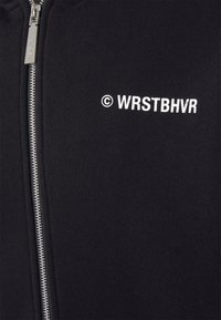 Hoodie noir zippé en tissu coton lisse, avec le texte imprimé en blanc "© WRSTBHVR" sur le côté gauche et une fermeture éclair en métal.
