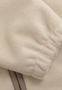 Cream-colored fleece fabric with a soft texture, featuring a gathered cuff and contrasting brown zipper accents.