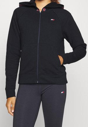 Mujer con sudadera negra con cremallera y leggings negros, ambos con pequeños logotipos rectangulares en rojo, blanco y azul.