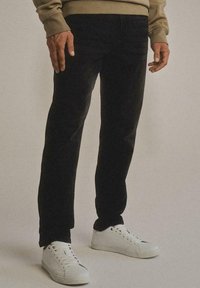 Uomo che indossa pantaloni neri, sneakers bianche e una maglietta beige a maniche lunghe, in piedi davanti a uno sfondo semplice.