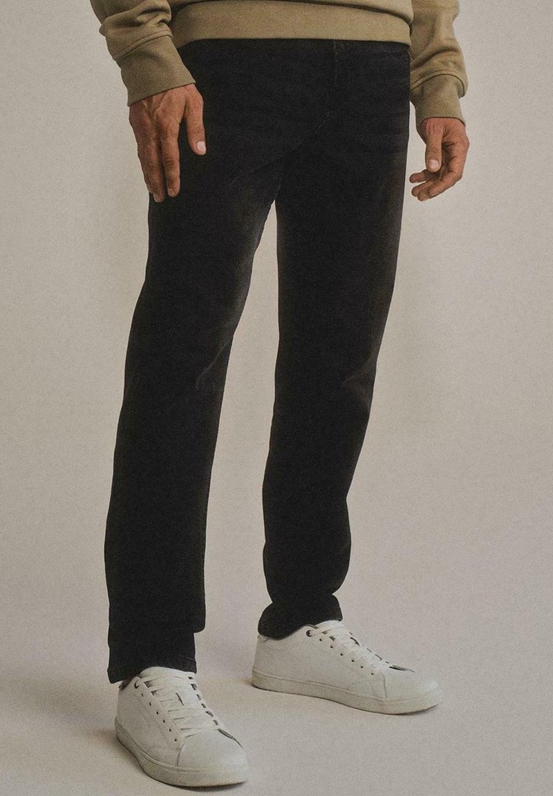 Uomo che indossa pantaloni neri, sneakers bianche e una maglietta beige a maniche lunghe, in piedi davanti a uno sfondo semplice.
