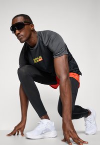 Mandligt atletisk outfit bestående af en sort kortærmet top med gul "BERLIN RUN" tekst, sorte leggings og hvide løbesko.