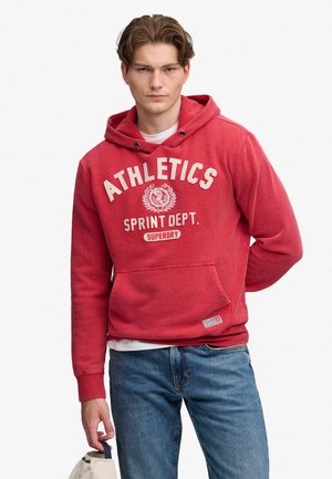 Jonge man met een rode hoodie met de tekst "ATHLETICS SPRINT DEPT. SUPERDRY", blauwe spijkerbroek, houdt een beige pet in zijn linkerhand.