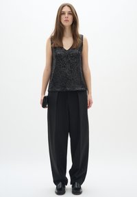 Top nero senza maniche con una superficie testurizzata, abbinato a pantaloni larghi neri. La modella tiene una piccola pochette nera e indossa scarpe nere.