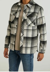 Ruudullinen flanellipaita musta-valkoisena, jossa on kaksi rintataskua, nappisulku ja hieman tekstuurinen pinta. Parin päällä beige t-paita.