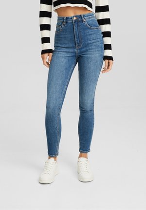 Woman wearing high-waist blue skinny jeans, white sneakers, and a black-and-white striped cropped sweater against a plain background.