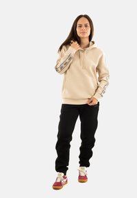 Felpa beige con accenti neri del logo sulle maniche, dotata di una tasca frontale. Abbinata a pantaloni della tuta neri e sneaker multicolori.