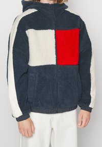 Fleece zip-up jakke med marineblå og cremefarvede paneler samt en rød kvadratisk detalje. Har hætte, elastiske manchetter og lommer. Blødt, tekstureret stof.