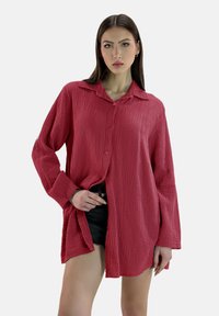 Camicia rossa a maniche lunghe con una texture, con design a bottone, vestibilità rilassata e colletto. Indossata sopra pantaloni corti neri, che mostrano uno stile casual.