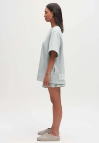 Lichtblauw oversized t-shirt met korte mouwen, zachte textuur en een logo-detail aan de onderkant, gecombineerd met grijze instappers en shorts.