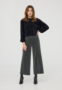 Maglione corto nero e peloso abbinato a pantaloni grigi a gamba larga a vita alta con texture. Il modello indossa stivaletti neri alla caviglia. Sfondo semplice.