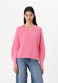 Rosa strikkegenser med vid rund hals, ribbestruktur og senkede skuldre, parret med blå denimjeans.