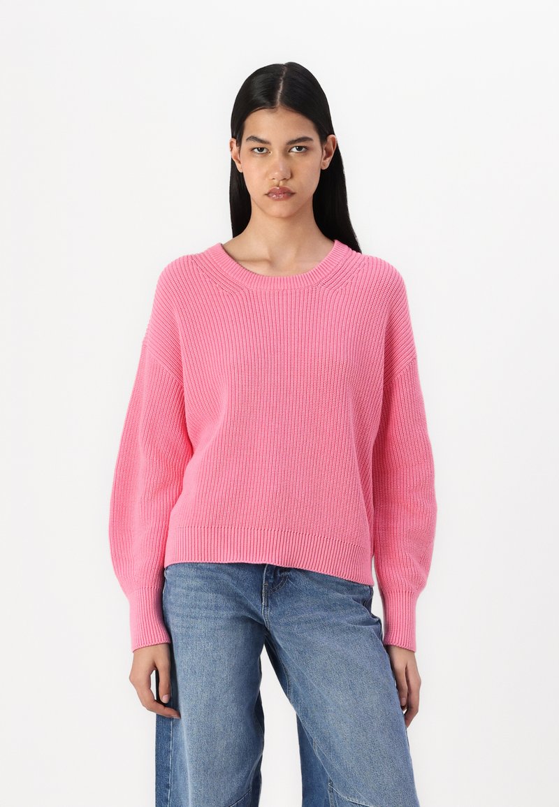Rosa strikkegenser med vid rund hals, ribbestruktur og senkede skuldre, parret med blå denimjeans.