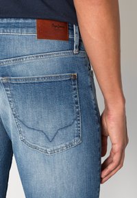 Light blue denim jeans with a subtle fade, featuring a brown leather brand tag, back pockets with stitching detail, and a slim fit.