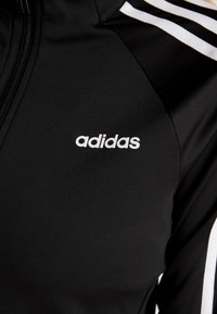 Černá sportovní bunda s bílým logem adidas. Má zipové zapínání a tři bílé pruhy na ramenou. Hladká a elastická tkanina.