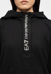 Άτομο που φοράει μαύρο φούτερ με κουκούλα με λευκό κάθετο λογότυπο EA7 Emporio Armani στο κεντρικό εμπρόσθιο πάνελ.